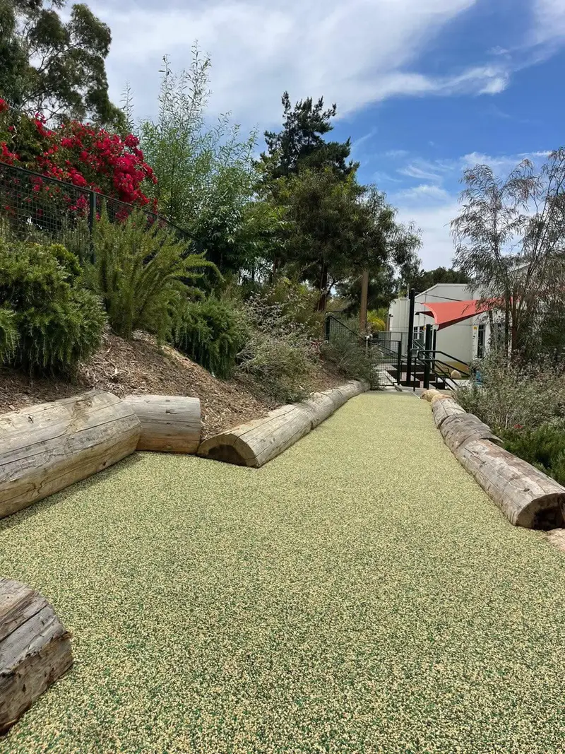 Smooth tan and green poured in place rubber pathway bordered by wooden logs and landscaped slopes at a San Diego school, installed by Turbo Surfacing Solutions